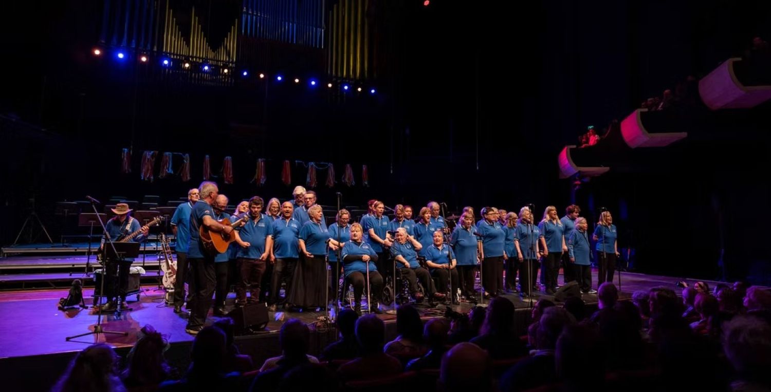 Large community choir wearing matching blue shirts performs on a brightly lit theatre stage. An acoustic guitarist accompanies them on the left, while a seated audience watches from the dim auditorium
